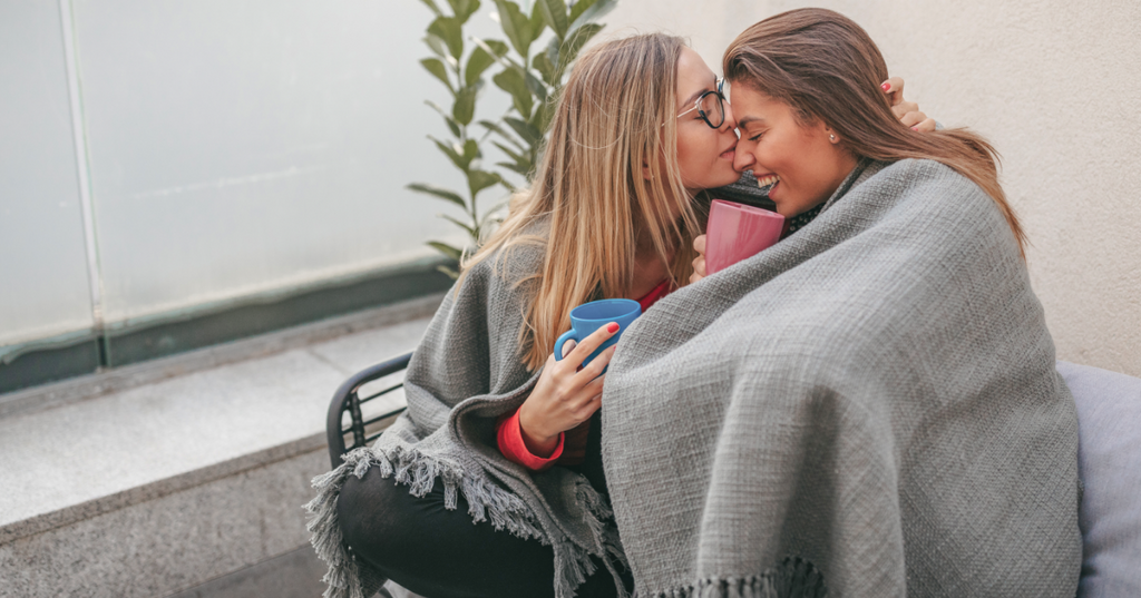 Two women sharing a blanket and holding mugs sneak a nose kiss.