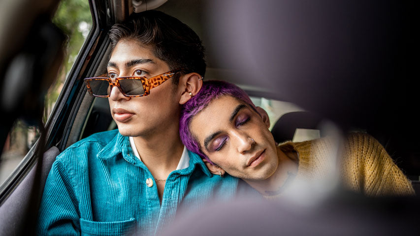 Person resting their sleepy head on their partner wearing sunglasses and looking out the car window.