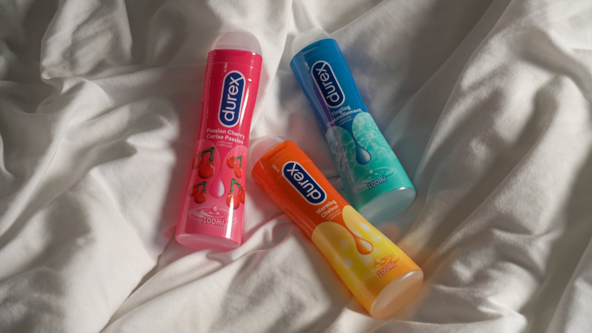 Bouteilles de lubrifiant Durex Cerise Passion, de lubrifiant Durex Chatouillement et de lubrifiant Durex Chaleur disposées sur un drap blanc. 