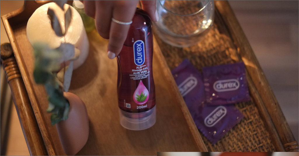 Une main qui s’étire vers la bouteille de lubrifiant Play Massage 2 en 1 Aloe Vera de Durex, à côté de condoms Durex sur une table de nuit. 