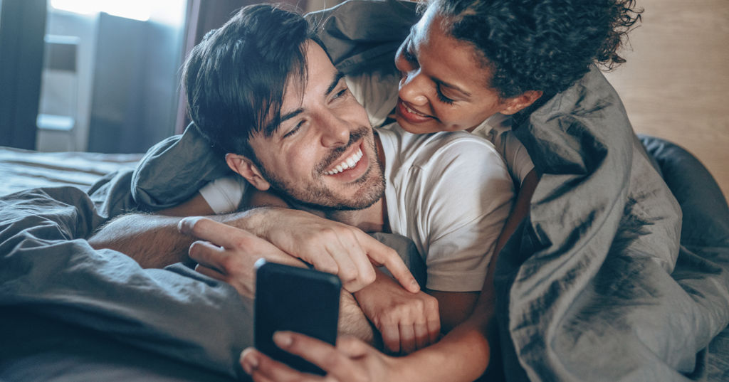 Un couple apprend à s’améliorer au lit en consultant un téléphone en riant. 