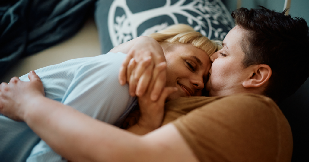 Two partners holding each other in bed while one kisses the other’s forehead.