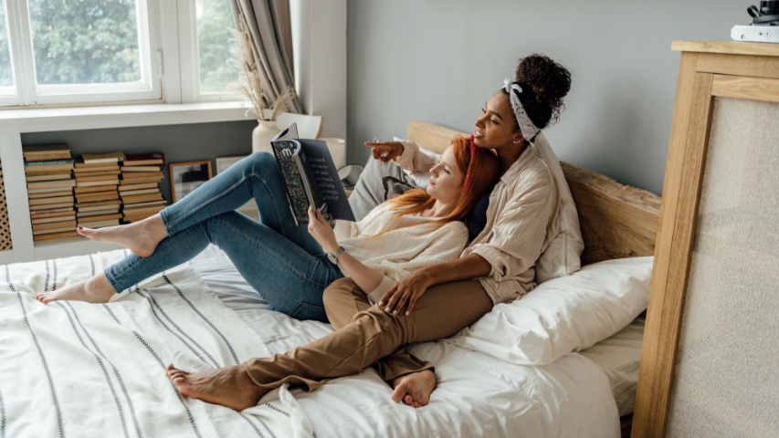 Two women reading a book together on allyship in bed.