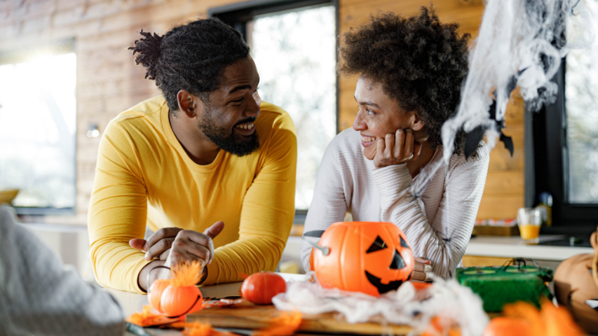 Woman and man smiling at each other while leaning on a table filled with Halloween decorations.
