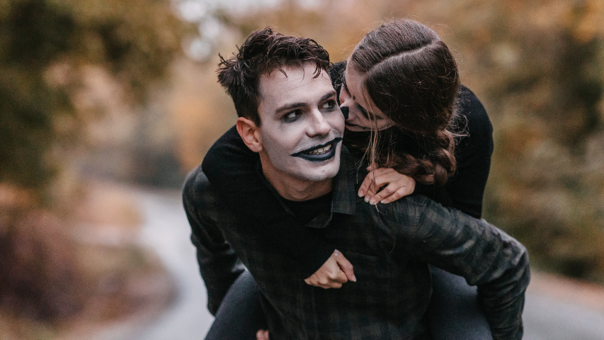 Man in white face paint and black lipstick carrying his partner on his shoulders outdoors on a Halloween holiday date.