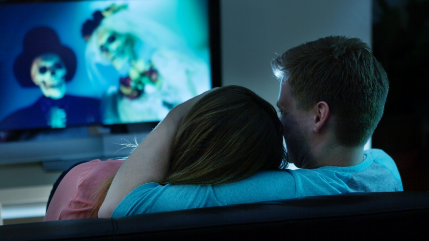Two people snuggling on the sofa and showing Halloween romance while watching a movie with a skeleton bride and groom.