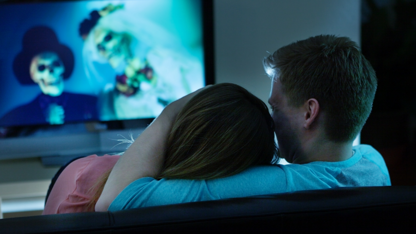 Deux personnes se blottissant sur le sofa et vivant une romance d’Halloween tout en regardant un film avec des mariés squelettiques.