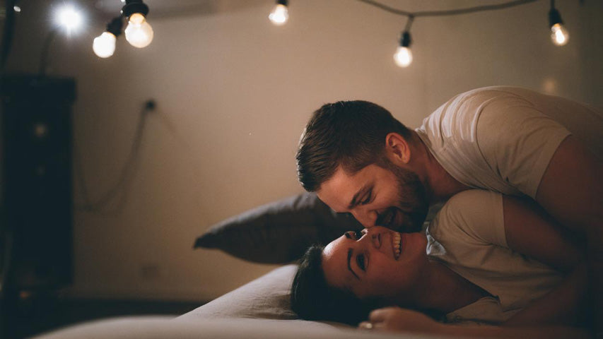 Dans un lit entouré de lumières suspendues, un homme embrasse la joue de sa partenaire qui sourit.
