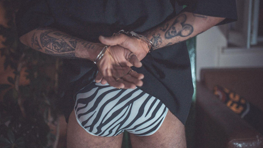 Tattooed man in zebra print briefs showing his handcuffed hands behind his back.
