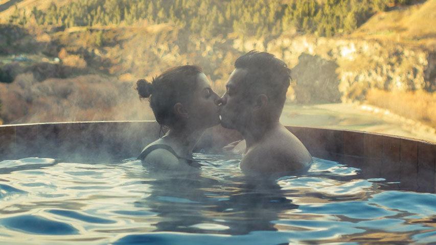 Two people kissing in a steamy hot tub across from a mountainous view.