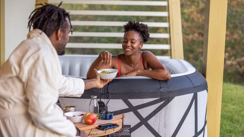 Man in robe feeding his girlfriend a bowl of fruit as she sits in a hot tub.