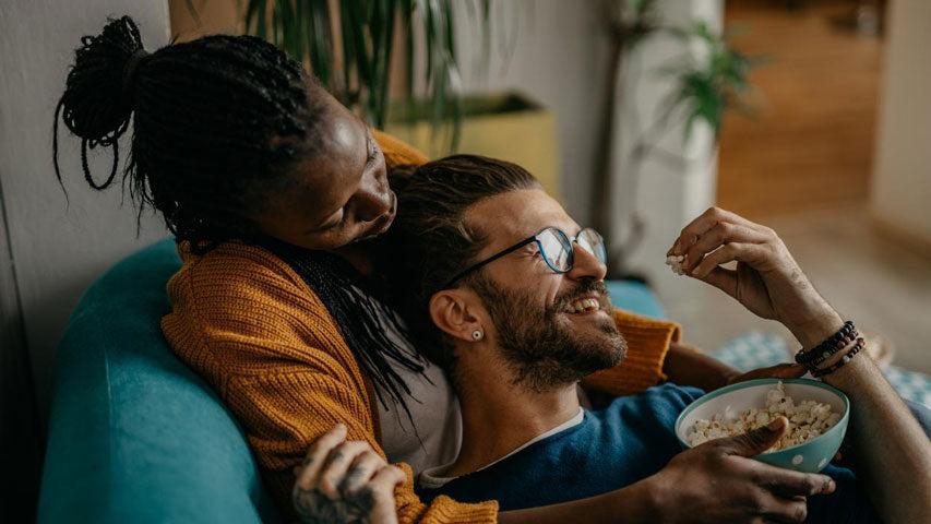 Une femme tient un bol de maïs soufflé pendant que son partenaire est étendu sur elle, en riant d’une idée pour la Saint-Valentin qu'ils ont eue en tête.