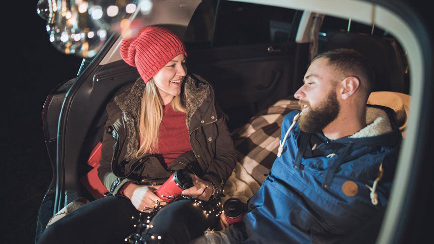 Woman and man wearing layers having a conversation with one another in the open trunk of their car next to hanging lights.