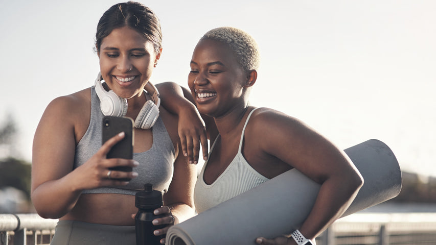 Une femme avec des écouteurs qui tient une bouteille d’eau noire et un téléphone en souriant alors que la femme à côté d’elle pose son bras sur son épaule et sourit en tenant un matelas de yoga sous le bras.