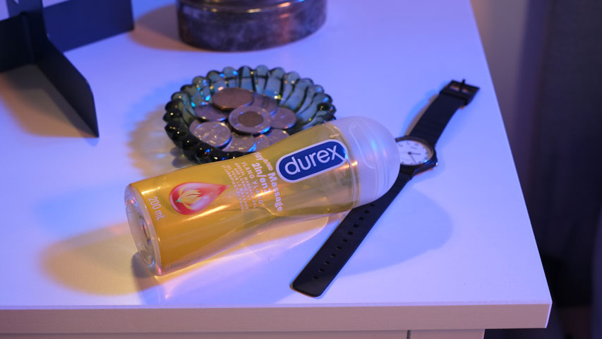 Sur une table de chevet, une bouteille de Durex Play Massage 2 en 1 Ylang Ylang est posée face contre table à côté d’une montre et d’un plateau à monnaie.