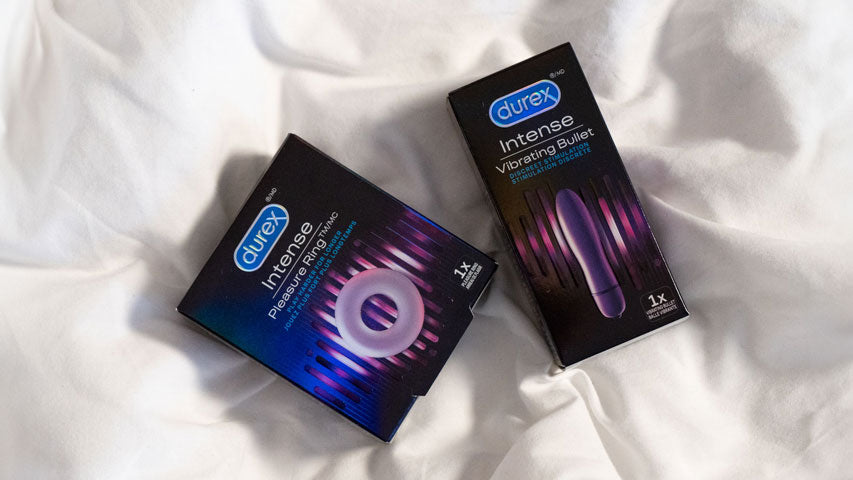 Les emballages d’un anneau pénien Durex Pleasure Ring et d’un vibromasseur Durex Intense sur un lit de draps blancs.