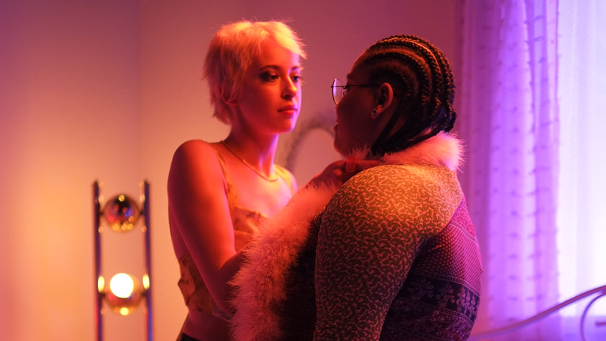 Two partners exploring a sensual massage with a feather boa amid purple and warm lighting.