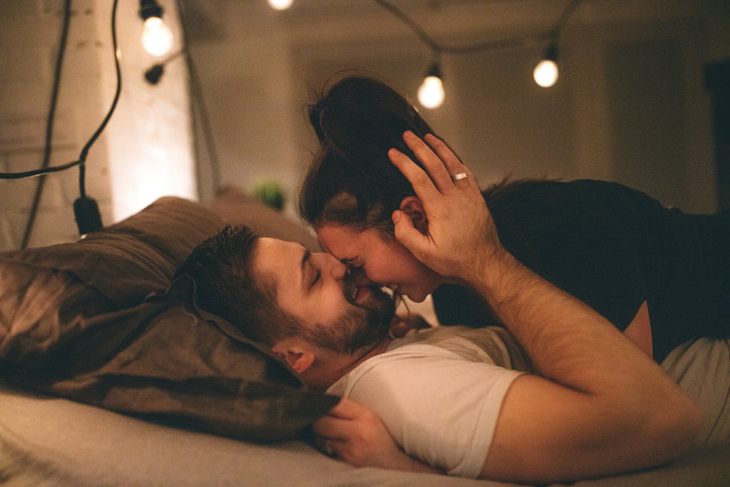 A woman lays on top of a man in bed while he kisses her nose.