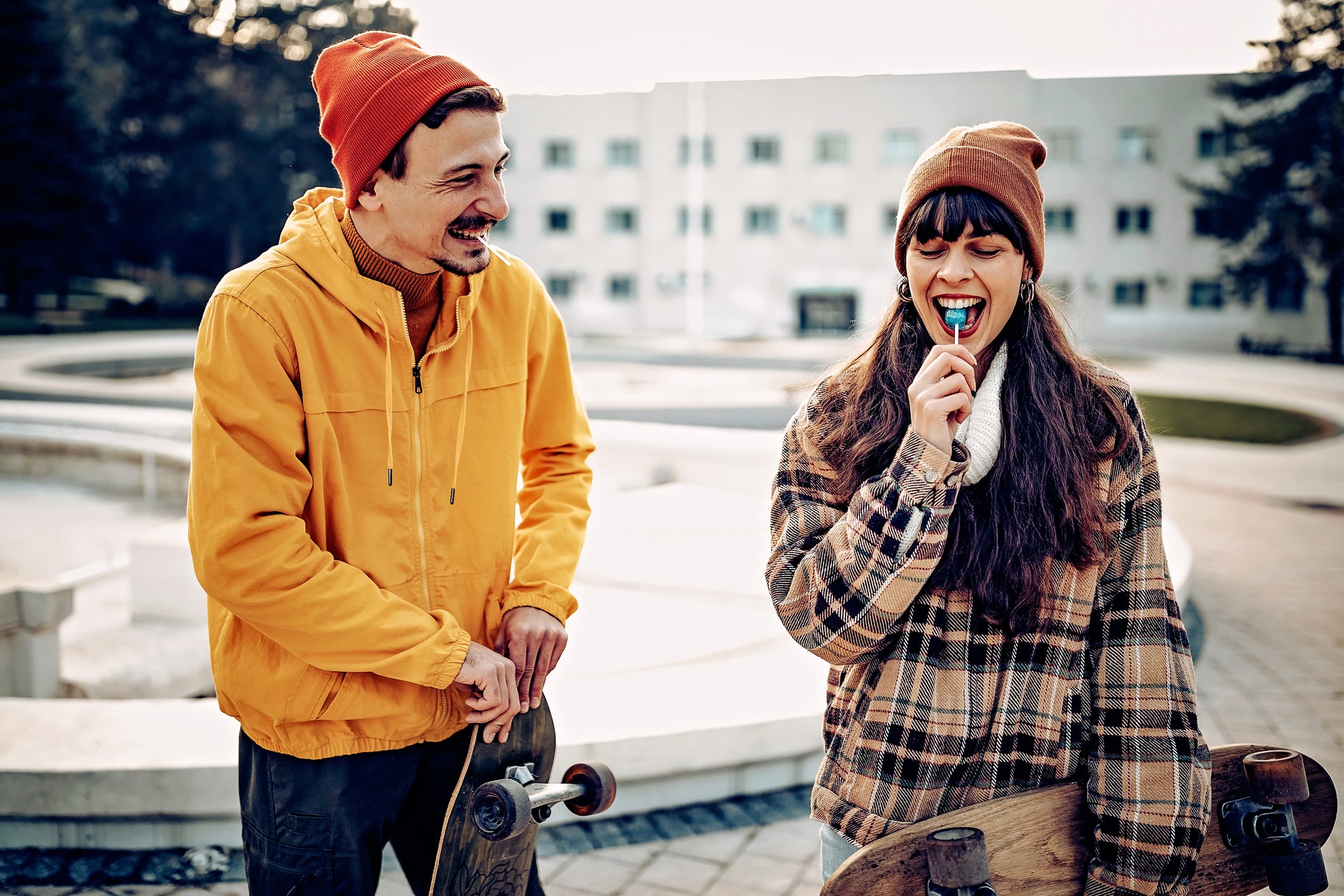 Un homme et une femme à l’extérieur qui tiennent leurs planches à roulettes et qui rient alors que la femme mange une sucette bleue.