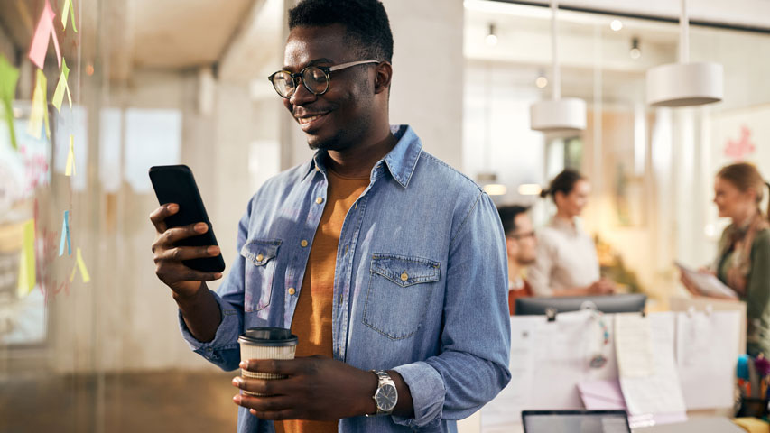 Un homme tient une tasse de café tout en souriant à son téléphone dans un espace de bureau.