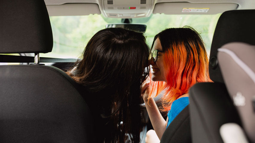Two women wearing glasses motioning for a kiss in the front seats of their car.