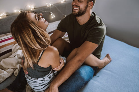 A couple smiles at each other on a bed while cuddling.