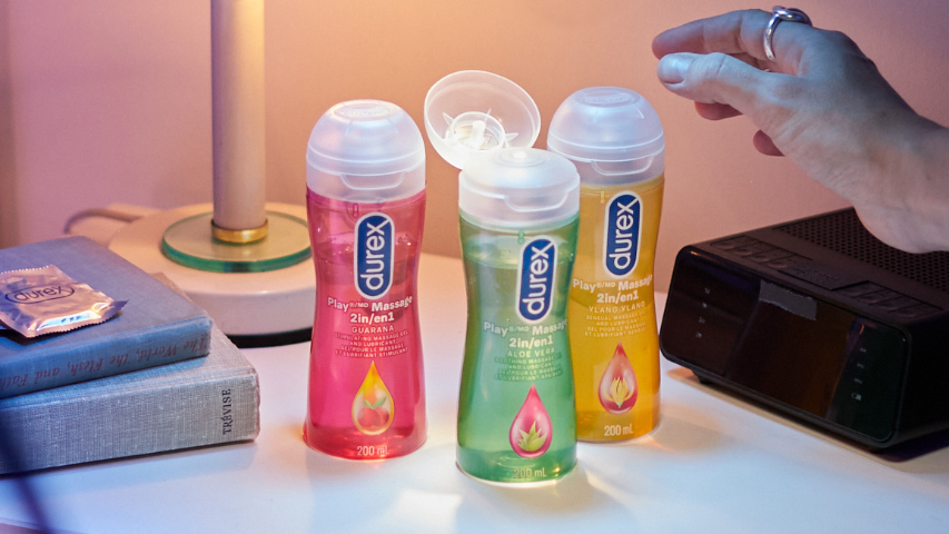 Hand reaching for an open bottle of Durex Play Massage 2 in 1 Aloe Vera next to Durex Play Massage 2 in 1 Guarana and Durex Play Massage 2 in 1 Ylang Ylang on a bedside table.