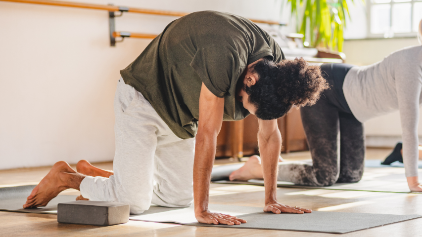 Un homme adopte la position de yoga du chat et de la vache tandis que sa partenaire fait de même à l’arrière.