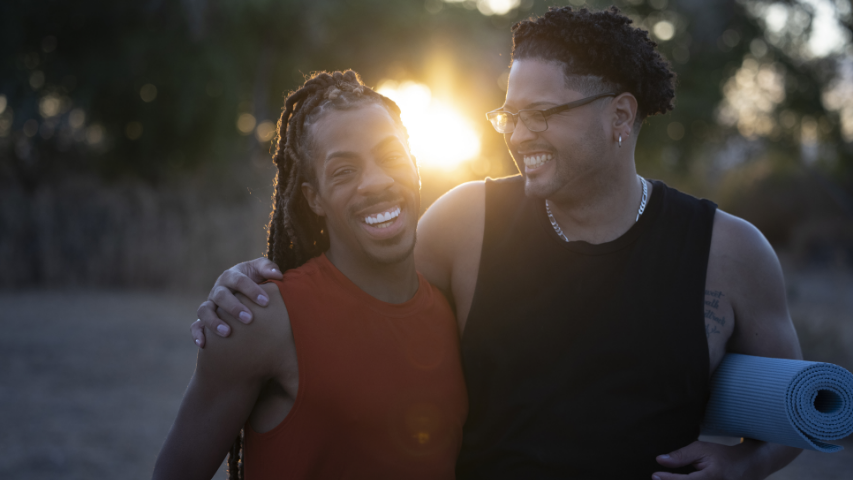 Two happy partners holding a yoga mat and embracing outdoors during sunset.