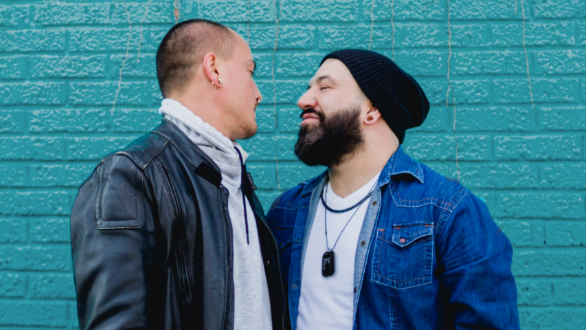 Deux hommes ferment les yeux et sont sur le point de s’embrasser devant des briques turquoises.