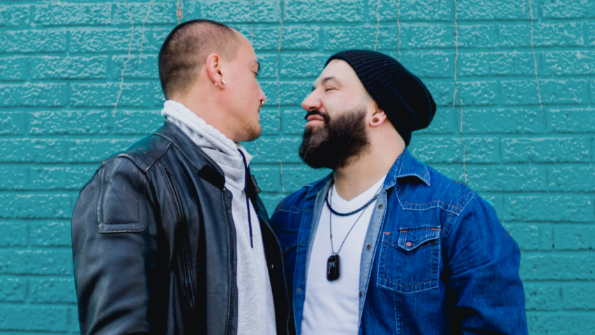 Two men closing their eyes about to go in for a kiss in front of teal brick.