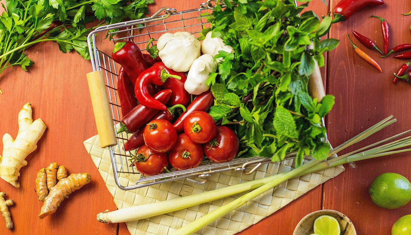 Basket of peppers, garlic, and greens sitting on top of a woven placemat with ginger root, limes, and other greens off to the side.