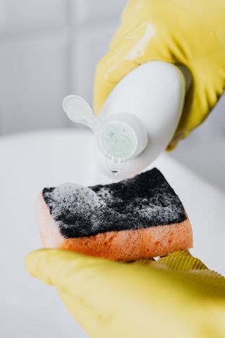 gloved hands holding a sponge and pouring a bottle of dish soap.