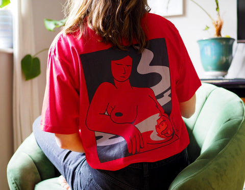 Red t-shirt with an illustration of a confident woman standing while containing a fire in her lower abdomen, with smoke dancing around her as the fire turns down. Illustration by artist Ana Curbelo, Untepid 2021.