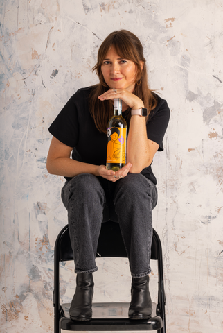 White woman (illustrator Ana Curbelo) sits on the back of a metallic chair in front of a grey and white background. Her head rests over her hands, which hold and frame a white wine bottle of Pinot Grigio. The bottle's label is bright orange and purple, with an illustration of a woman holding a flower looking directly at the viewer, illustrated by the artist holding it.