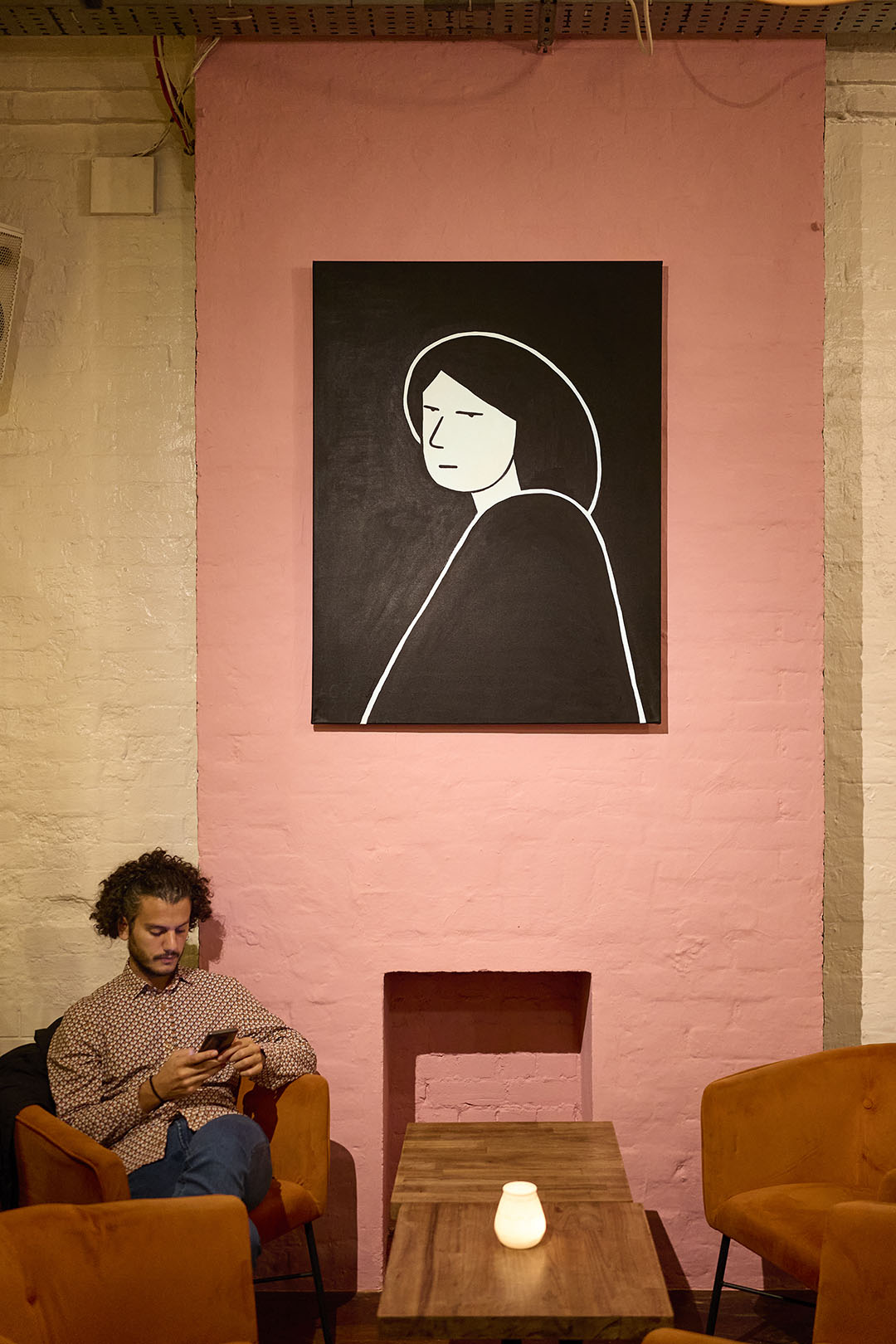 Oil painting of a black and white figure by artist Ana Curbelo is hung on a pink wall over a man sat underneath looking at his phone.