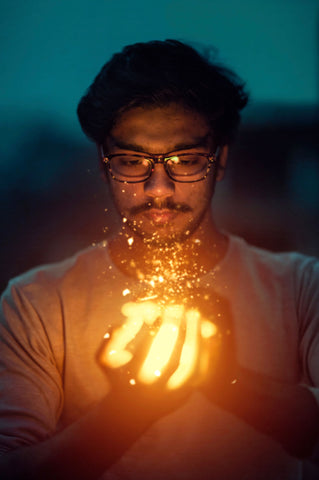 Man Holding a ball of light that looks like fire with embers sparking out of his hand, the man has olive skin, brown hair, is wearing glasses, and it looks like a night time sky in the background