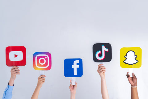 Five different hands holding up signs with the social media icons for youtube, instagram, facebook, tik tok and snapchat