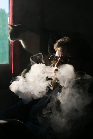 Guy smoking a vape battery with glasses on and a cat in the background