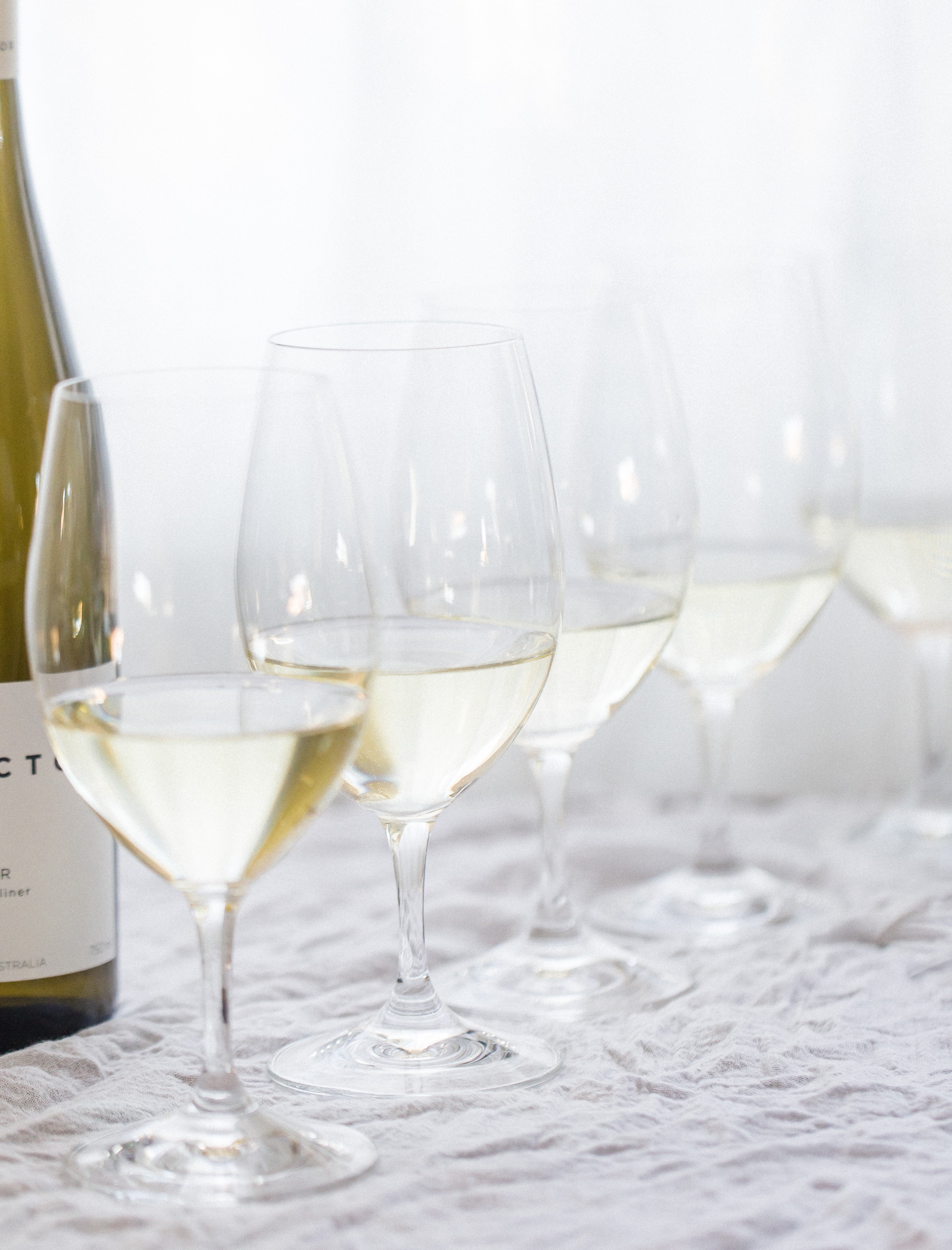 A row of four wine glasses with white wine in them, on a white linen tablecloth. A bottle of Collector Wines is just out of shot.