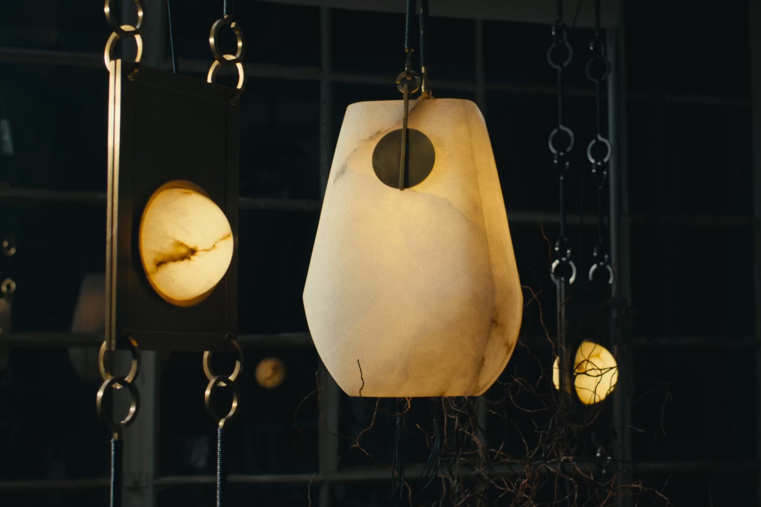 A white basket-shaped alabaster lantern with brass accents, suspended by leather-wrapped hooks, hangs in a darkened room.