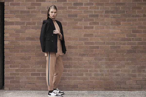 Image of a model wearing a matching cashmere tracksuit in camel colour, with a black blazer draped over her shoulders