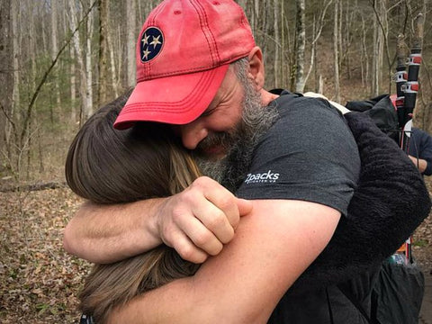 A man in a red hat hugging a brunette woman in the forest.