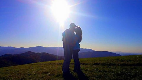 A couple kissing in a field with the sun shining in the background.