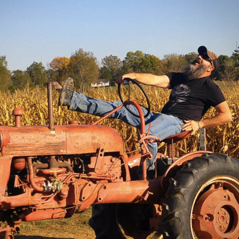 Bearded man leaning back and laughing on a tractor in a corn field.