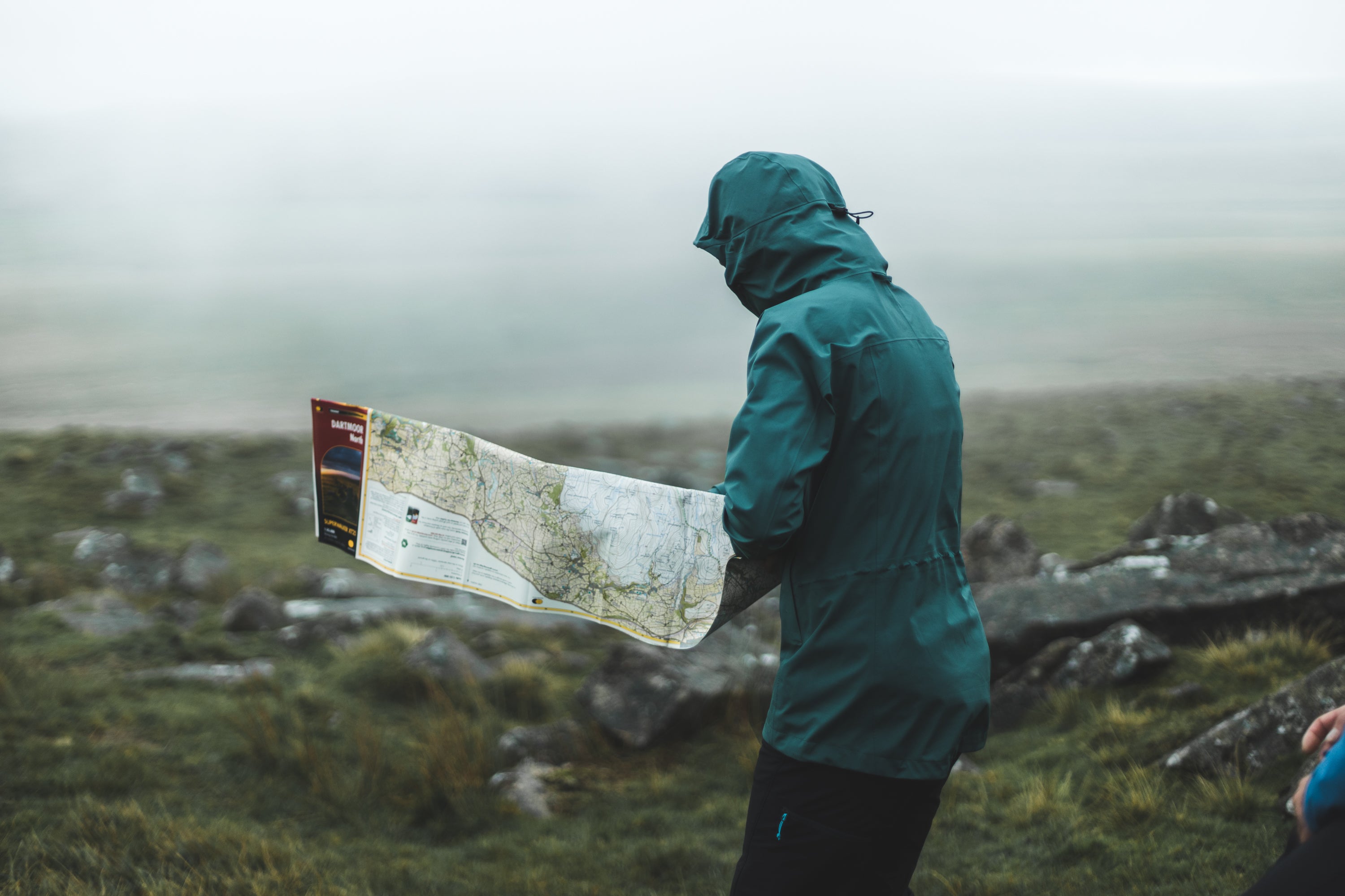 A woman using a Harvey Map on Dartmoor in strong winds, wearing a hillwalking waterproof jacket that's PFC free