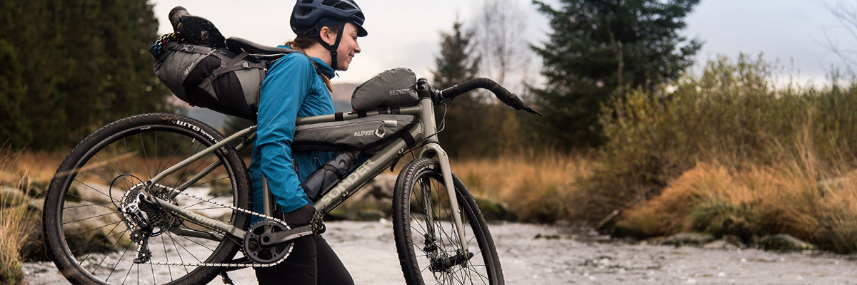 A woman carrying her bikepacking bike with grey bikepacking bags on through a river