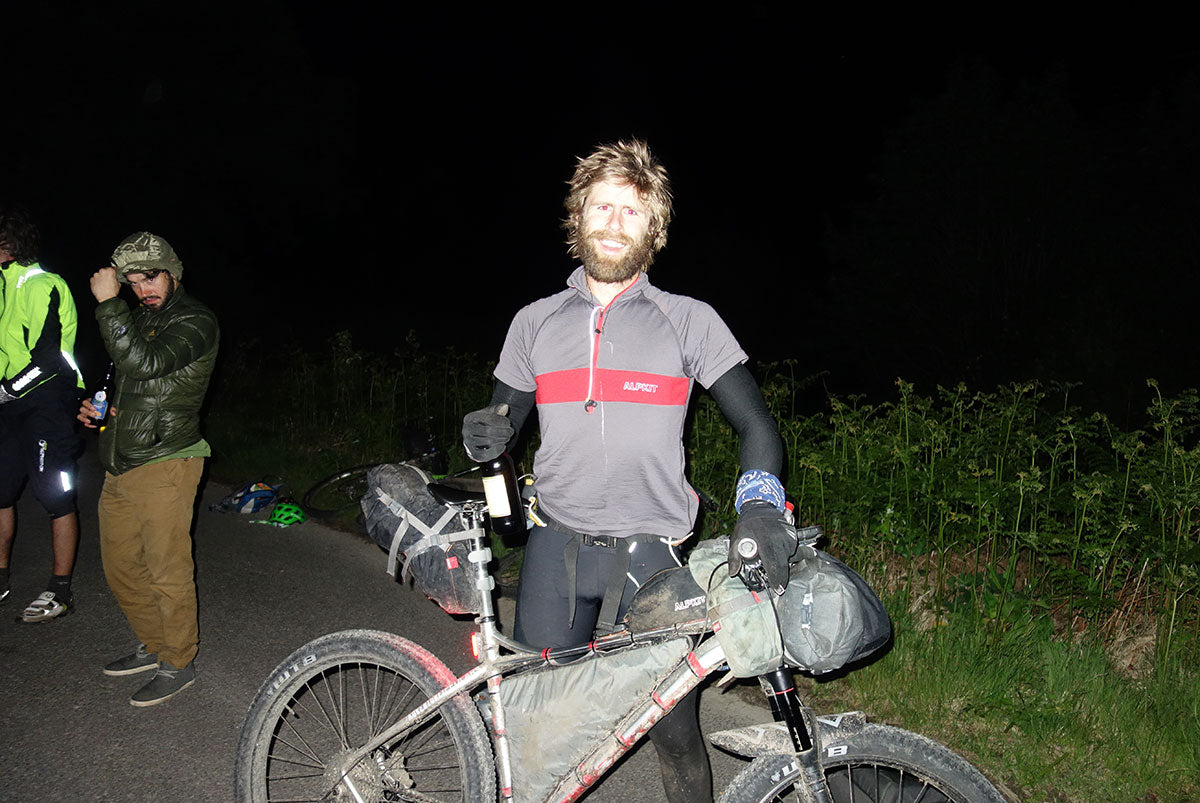 Pete at the finish line at night, his bike in one hand, and a bottle of wine in the other