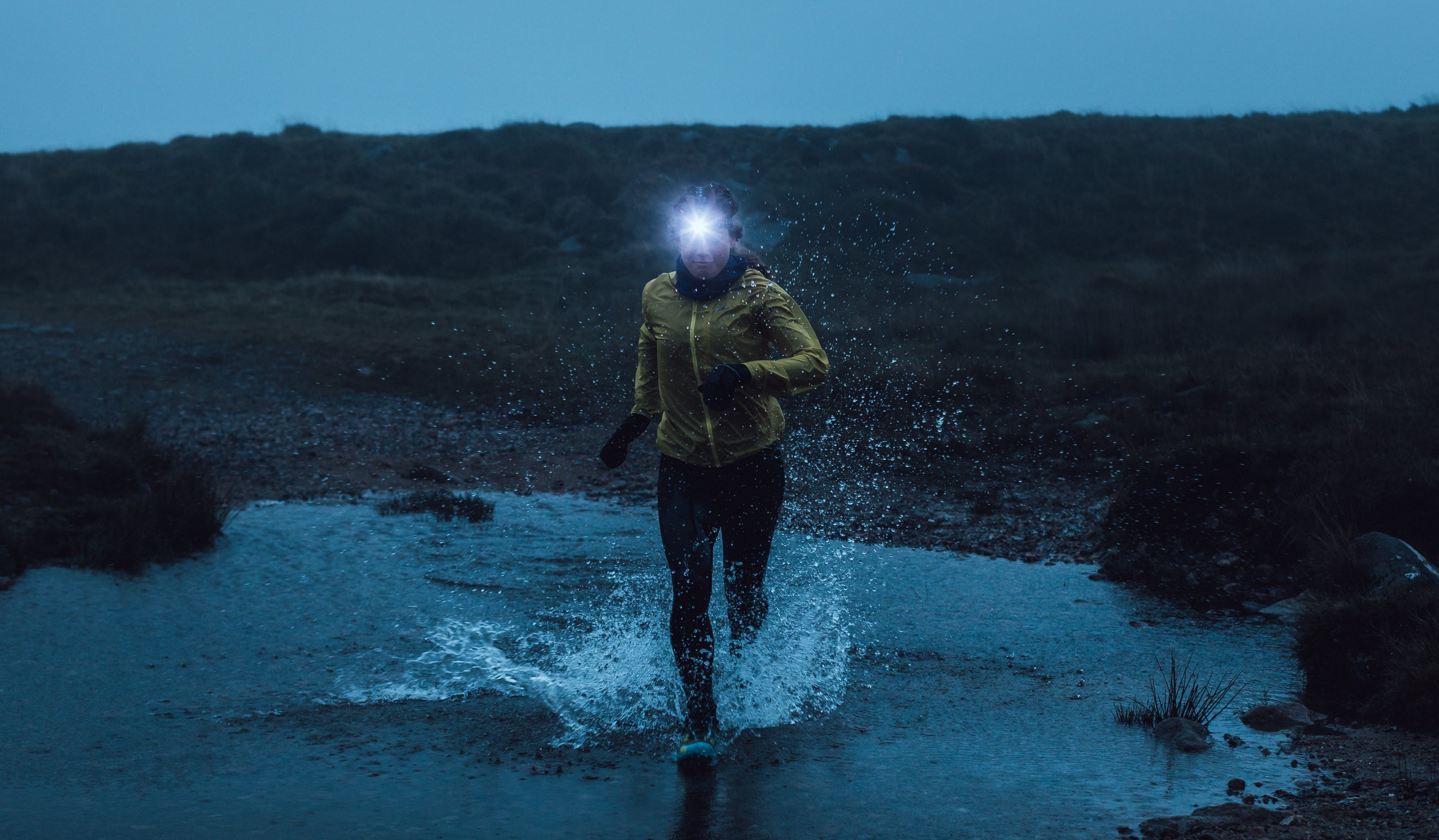 A woman trail running on Dartmoor by head torch and splashing through a puddle