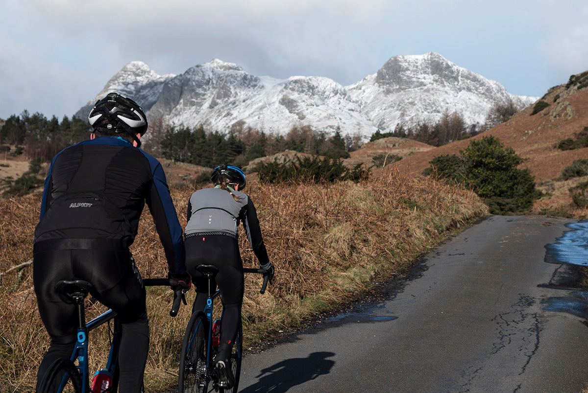 Two road cyclists climbing steep road against snowy backdrop